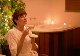 Mulher relaxando em um ambiente de spa, vestindo roupão branco e saboreando uma xícara de chá, com banheira ao fundo, luzes suaves e velas acesas, transmitindo paz e autocuidado.